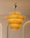A tiered glass chandelier with a warm golden-yellow shade and polished silver metal ceiling canopy, shown hanging in a beige interior setting.