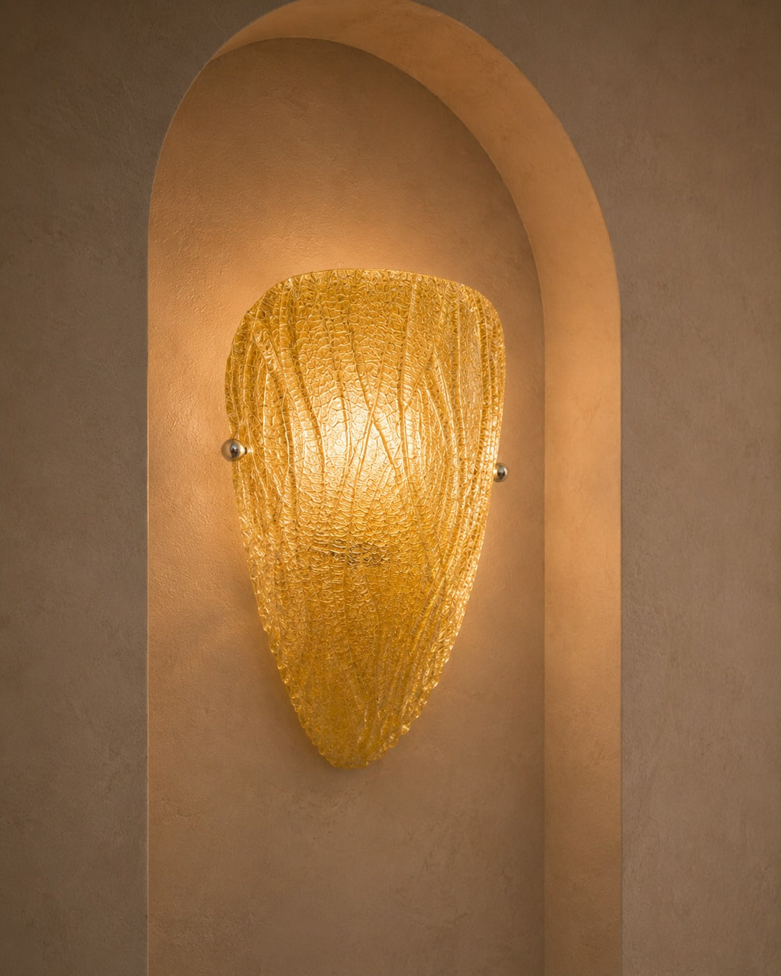 A warm golden textured glass sconce mounted on a beige plaster wall within a recessed archway.