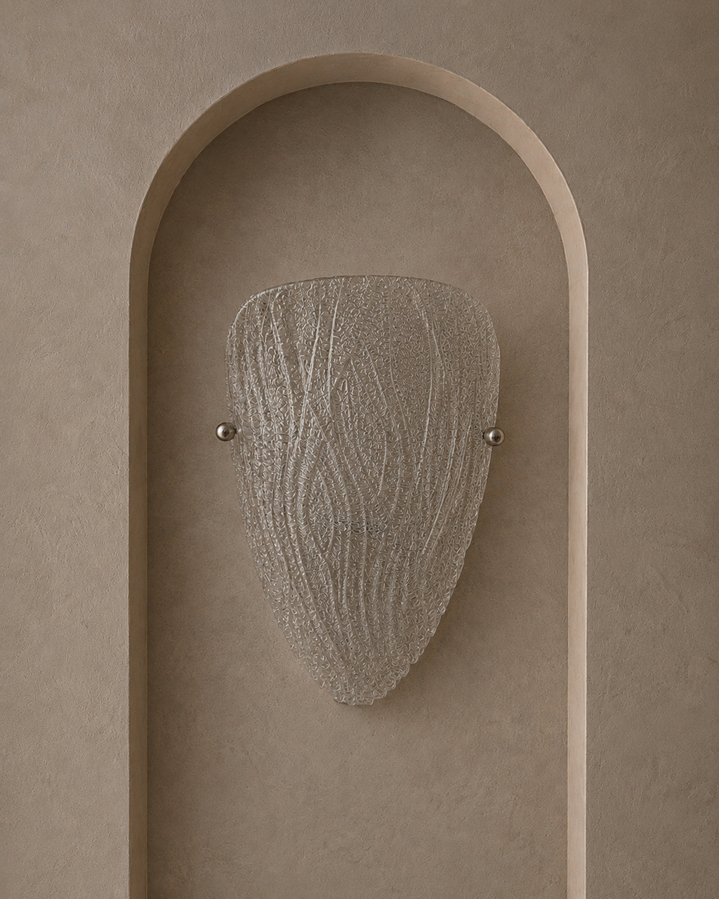 A modern wall sconce with a textured, organic-shaped frosted glass shade, mounted on a beige plaster wall with an arched recess.