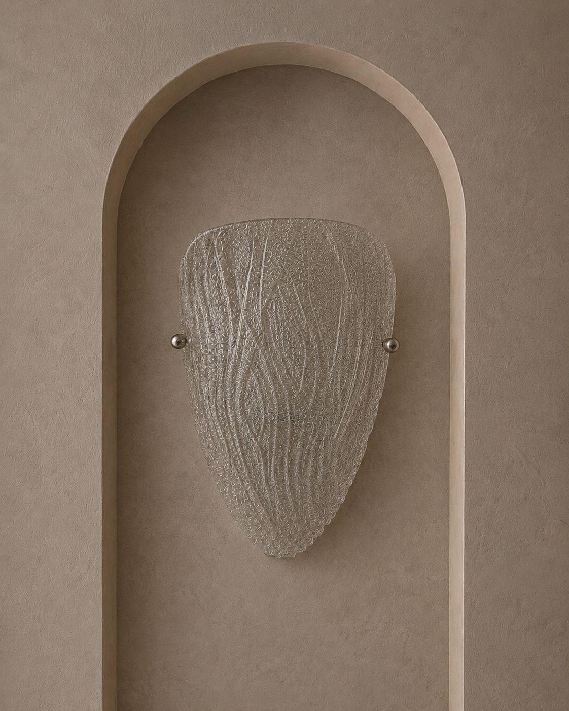 A modern wall sconce with a textured, organic-shaped frosted glass shade, mounted on a beige plaster wall with an arched recess.