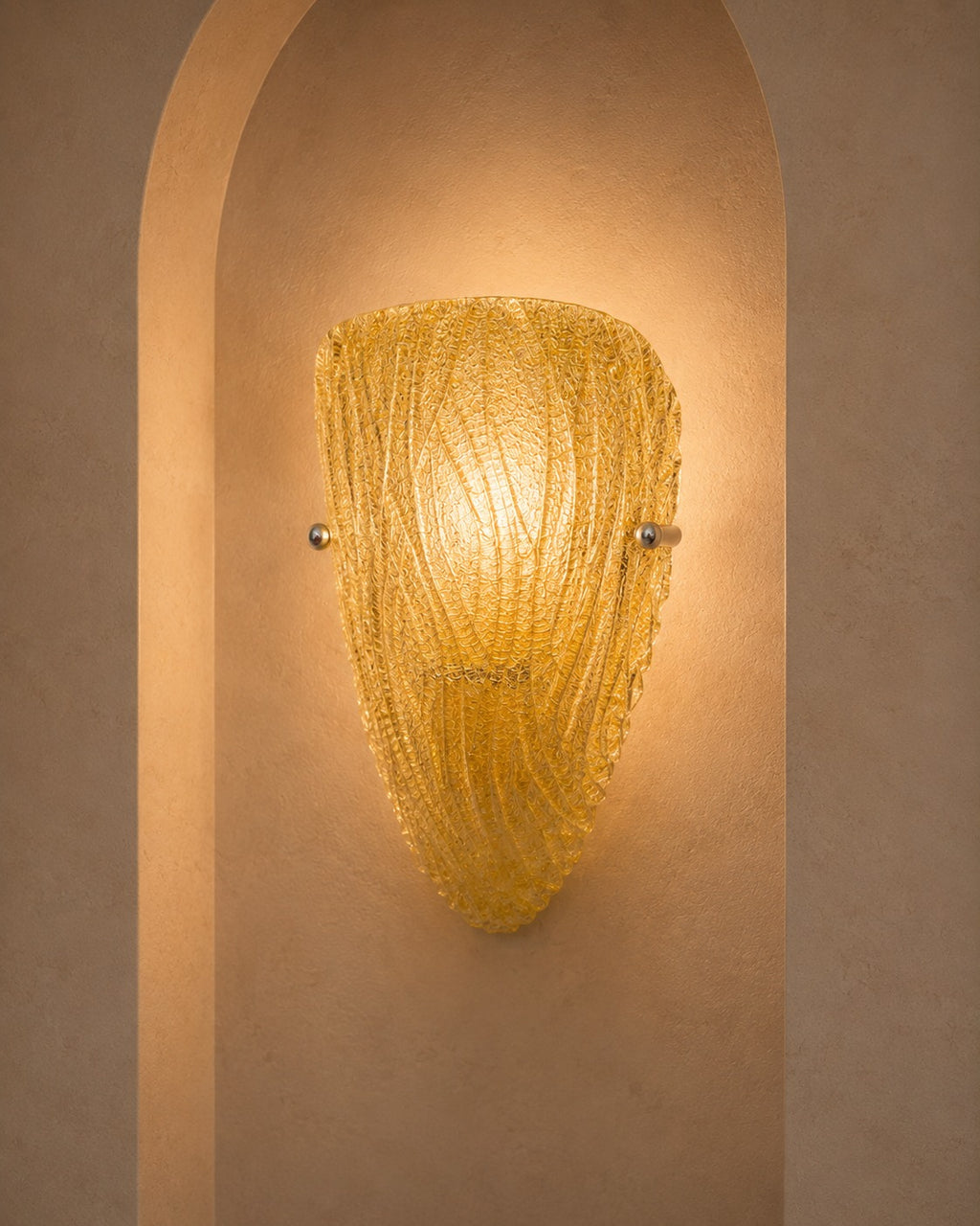 A textured, ribbed glass wall sconce in a warm golden-yellow hue, mounted on a beige plaster wall within a recessed archway.