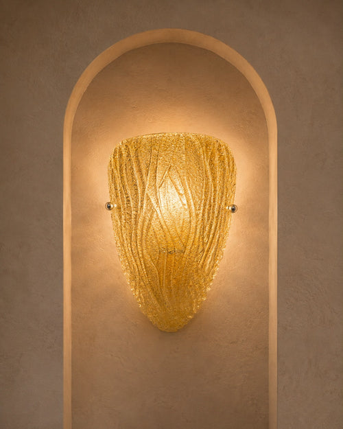 A textured golden-yellow glass sconce with a ribbed, organic shape, mounted on a warm beige plaster wall within an arched recess.
