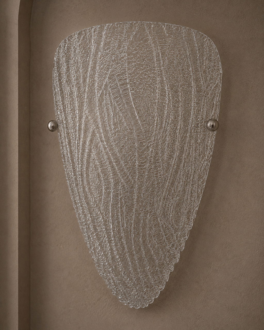 A modern wall sconce light fixture with a textured, organic-shaped clear glass shade and two small metallic silver mounting balls, mounted on a warm brown textured wall.