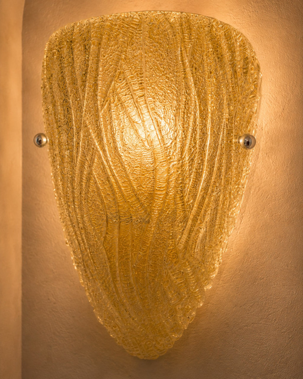 A textured gold glass wall sconce with a shield-like shape and wavy ribbed pattern, mounted on a warm beige plaster wall.