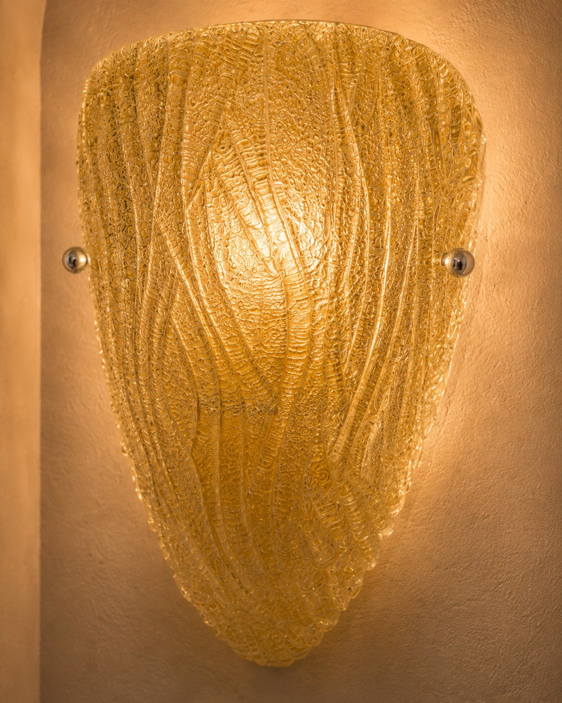 A textured gold glass wall sconce with a shield-like shape and wavy ribbed pattern, mounted on a warm beige plaster wall.