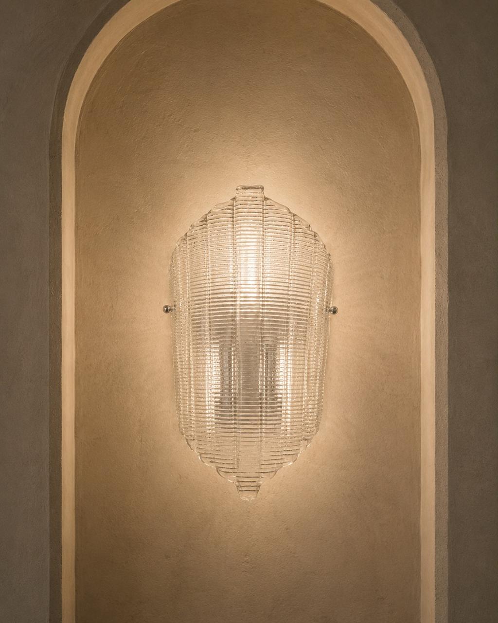 A modern ribbed glass wall sconce mounted on a warm beige plaster wall within an arched niche.