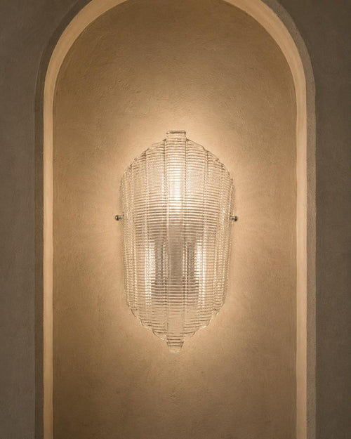 A modern ribbed glass wall sconce mounted on a warm beige plaster wall within an arched niche.