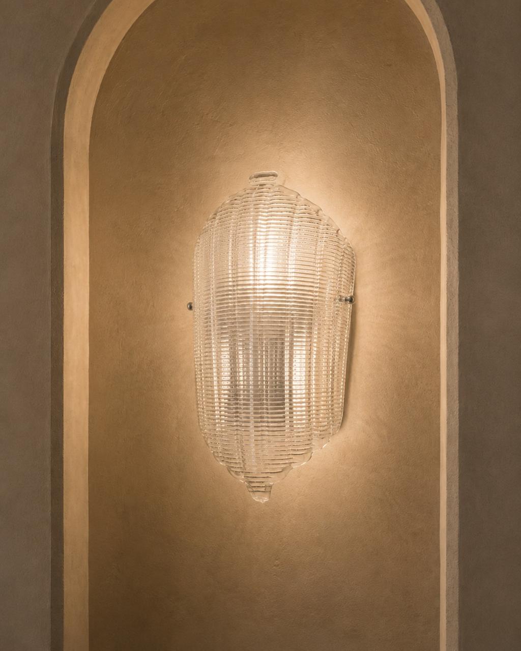 A clear ribbed glass wall sconce mounted on a warm beige plaster wall within an arched niche.