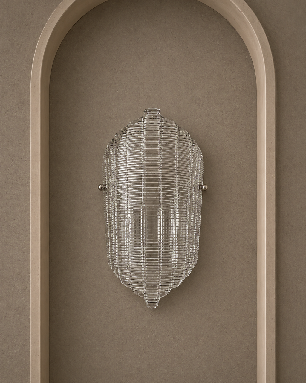 A modern ribbed glass wall sconce with a clear textured shade and small silver metal mounting hardware, mounted on a warm taupe wall within an arched recess.