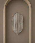 A modern ribbed glass wall sconce with a clear textured shade and small silver metal mounting hardware, mounted on a warm taupe wall within an arched recess.