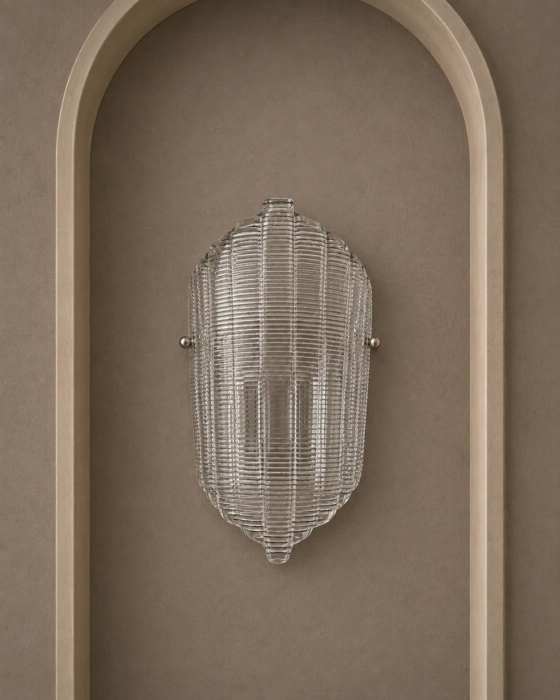 A modern ribbed glass wall sconce with a clear textured shade and small silver metal mounting hardware, mounted on a warm taupe wall within an arched recess.
