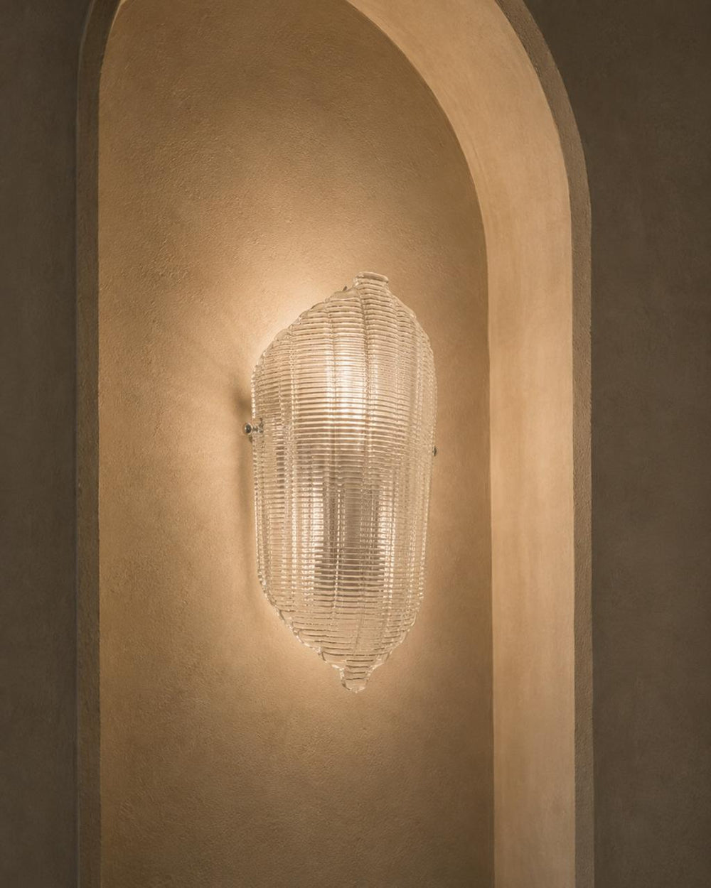 A modern ribbed clear glass wall sconce mounted on a warm beige plaster wall within an arched niche.