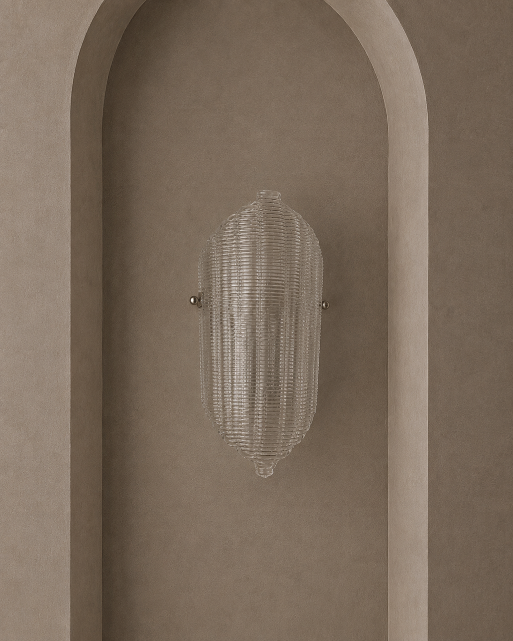 A modern wall sconce with a ribbed, textured clear glass shade and small metallic side hardware, mounted on a warm beige plaster wall with an arched recess.