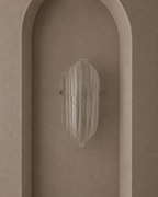 A modern wall sconce with a ribbed, textured clear glass shade and small metallic side hardware, mounted on a warm beige plaster wall with an arched recess.
