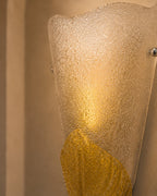 A close-up of a sculptural wall sconce with a textured frosted glass shade and a warm gold lower section, mounted against a beige wall.
