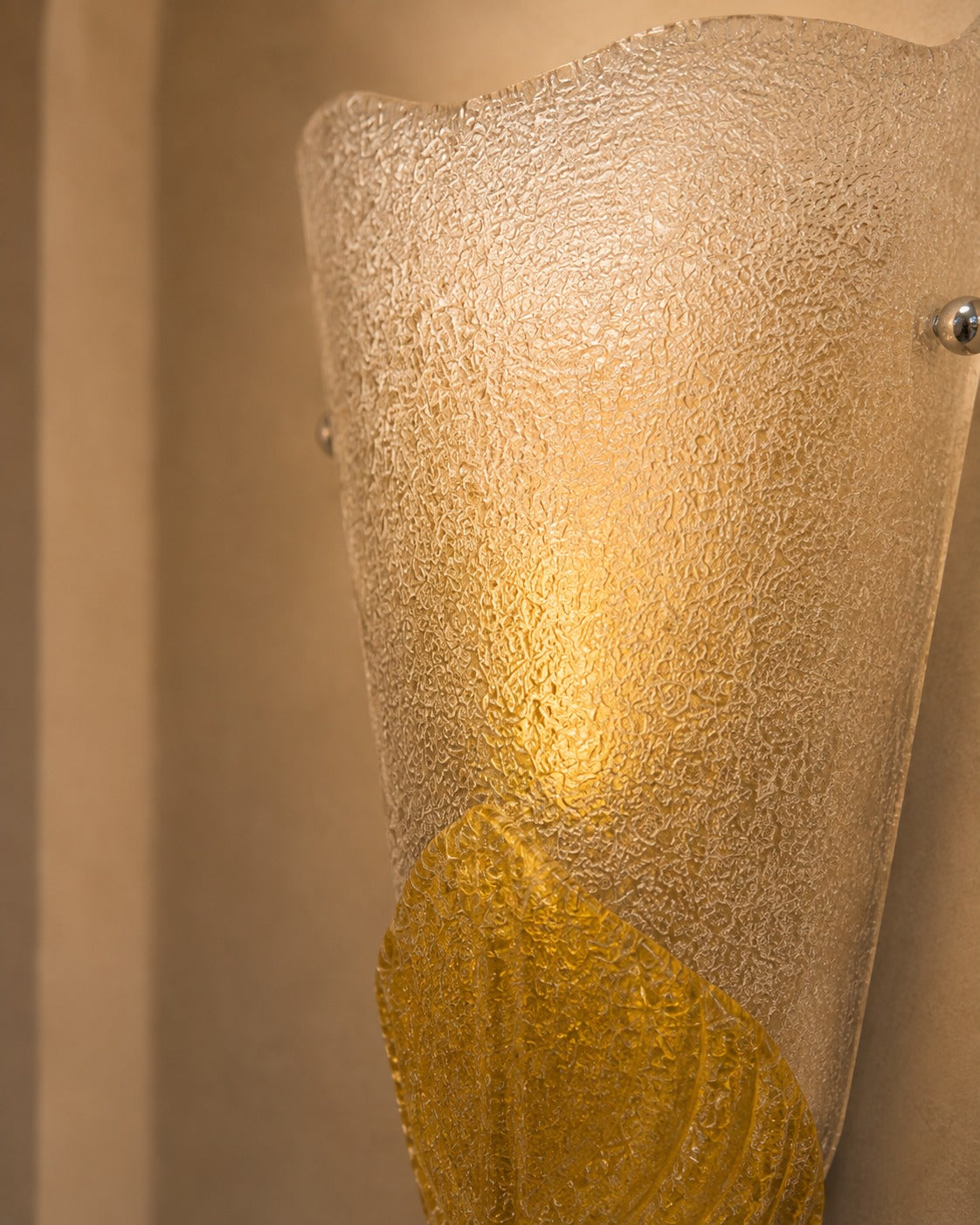 A close-up of a sculptural wall sconce with a textured frosted glass shade and a warm gold lower section, mounted against a beige wall.