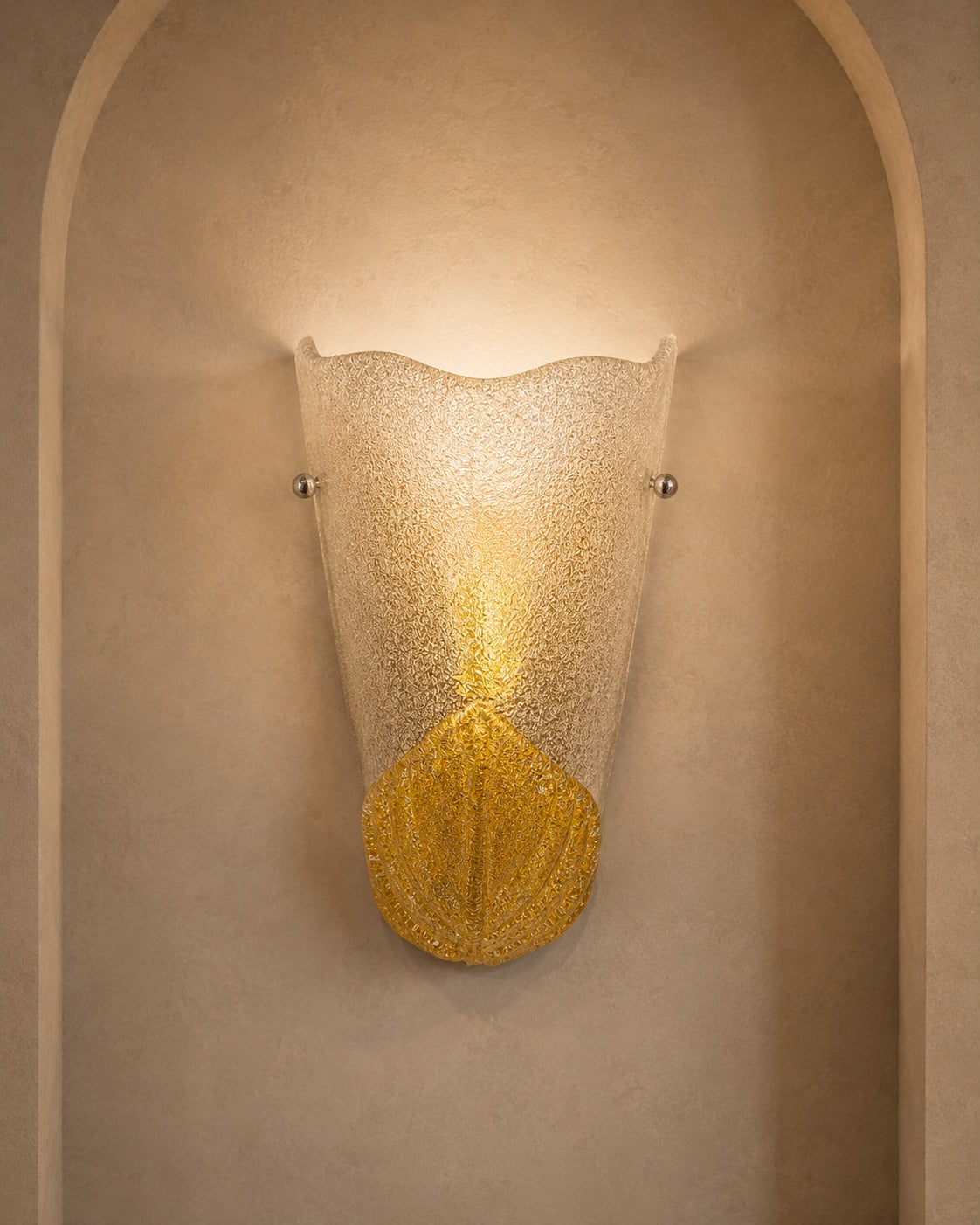 A sculptural wall sconce with a textured, scalloped frosted glass shade and a gold-toned lower section, mounted on a beige plaster wall within an arched niche.