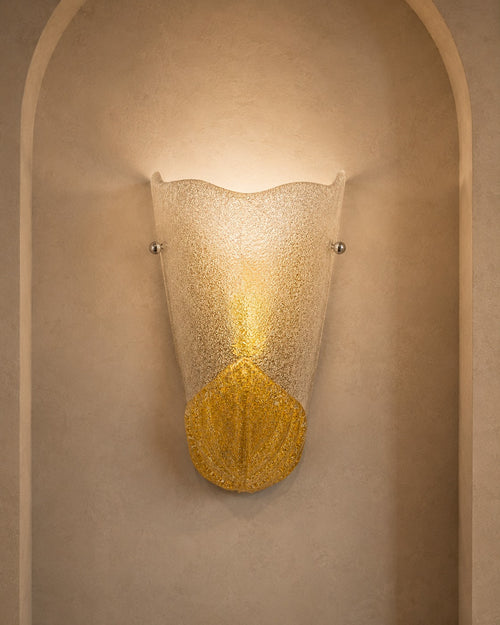 A sculptural wall sconce with a textured, scalloped frosted glass shade and a gold-toned lower section, mounted on a beige plaster wall within an arched niche.