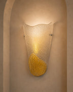 A modern wall sconce light fixture with a textured, frosted glass shade and a metallic gold leaf accent at the bottom, mounted on a beige plaster wall with an arched niche.