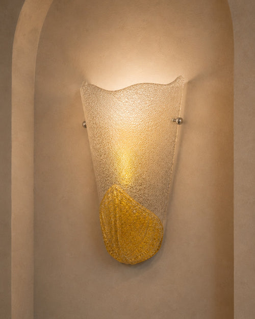 A modern wall sconce light fixture with a textured, frosted glass shade and a metallic gold leaf accent at the bottom, mounted on a beige plaster wall with an arched niche.