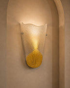 A modern wall sconce light fixture with a textured, translucent wavy glass shade and a metallic gold lower section, mounted on a beige stucco wall in an arched niche.