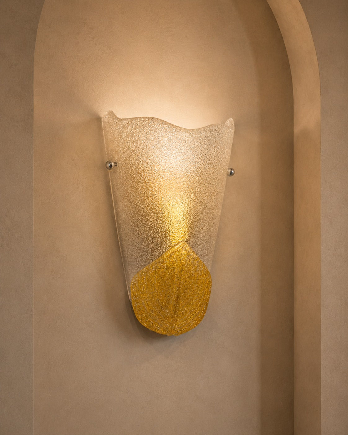 A modern wall sconce light fixture with a textured, translucent wavy glass shade and a metallic gold lower section, mounted on a beige stucco wall in an arched niche.