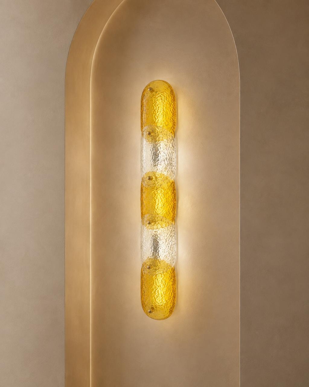 A modern vertical wall sconce with three textured amber-yellow glass bulbs set in a clear ribbed glass column, mounted on a warm beige arched wall.