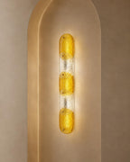 A modern vertical wall sconce with three textured amber-yellow glass bulbs set in a clear ribbed glass column, mounted on a warm beige arched wall.