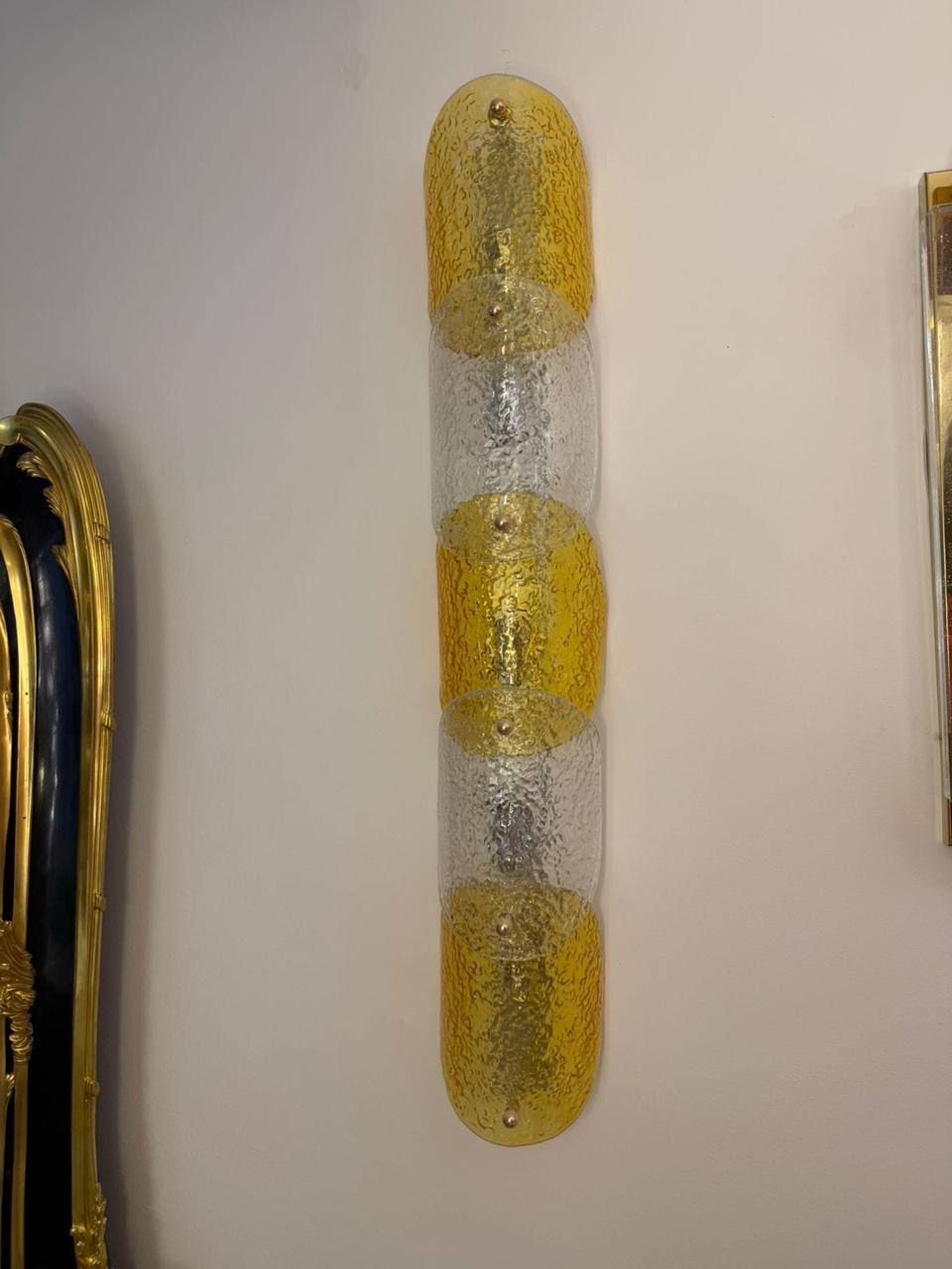 A vertical wall sconce with three textured gold glass sections alternating with clear textured glass sections, mounted on a plain off-white wall.