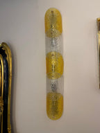 A vertical wall sconce with three textured gold glass sections alternating with clear textured glass sections, mounted on a plain off-white wall.