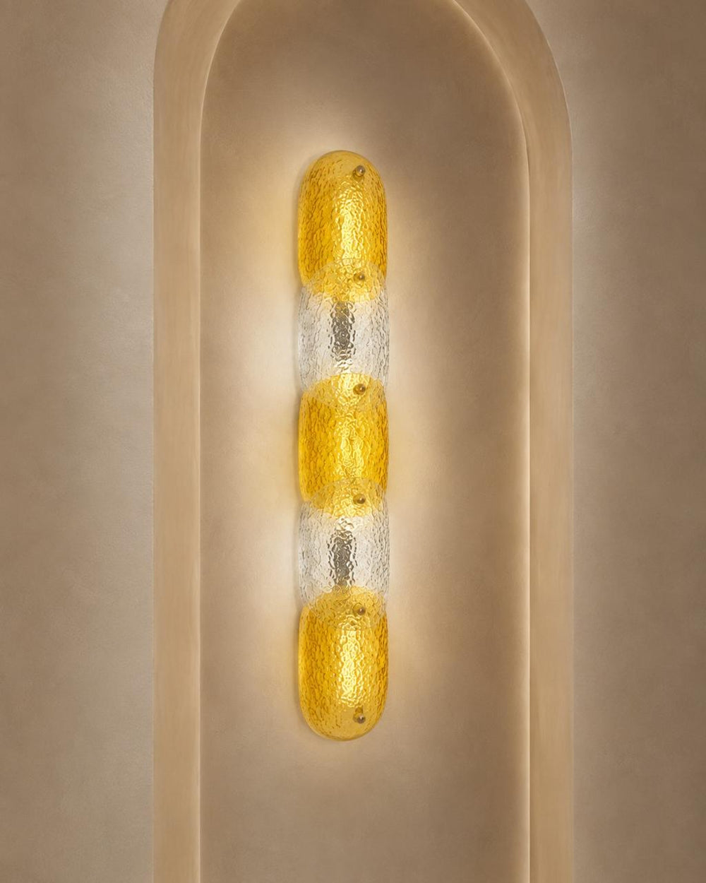 A modern vertical wall sconce with three textured yellow glass oval bulbs set in a clear glass column, mounted on a beige plaster wall with an arched recess.
