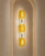 A modern vertical wall sconce with three textured yellow glass oval bulbs set in a clear glass column, mounted on a beige plaster wall with an arched recess.