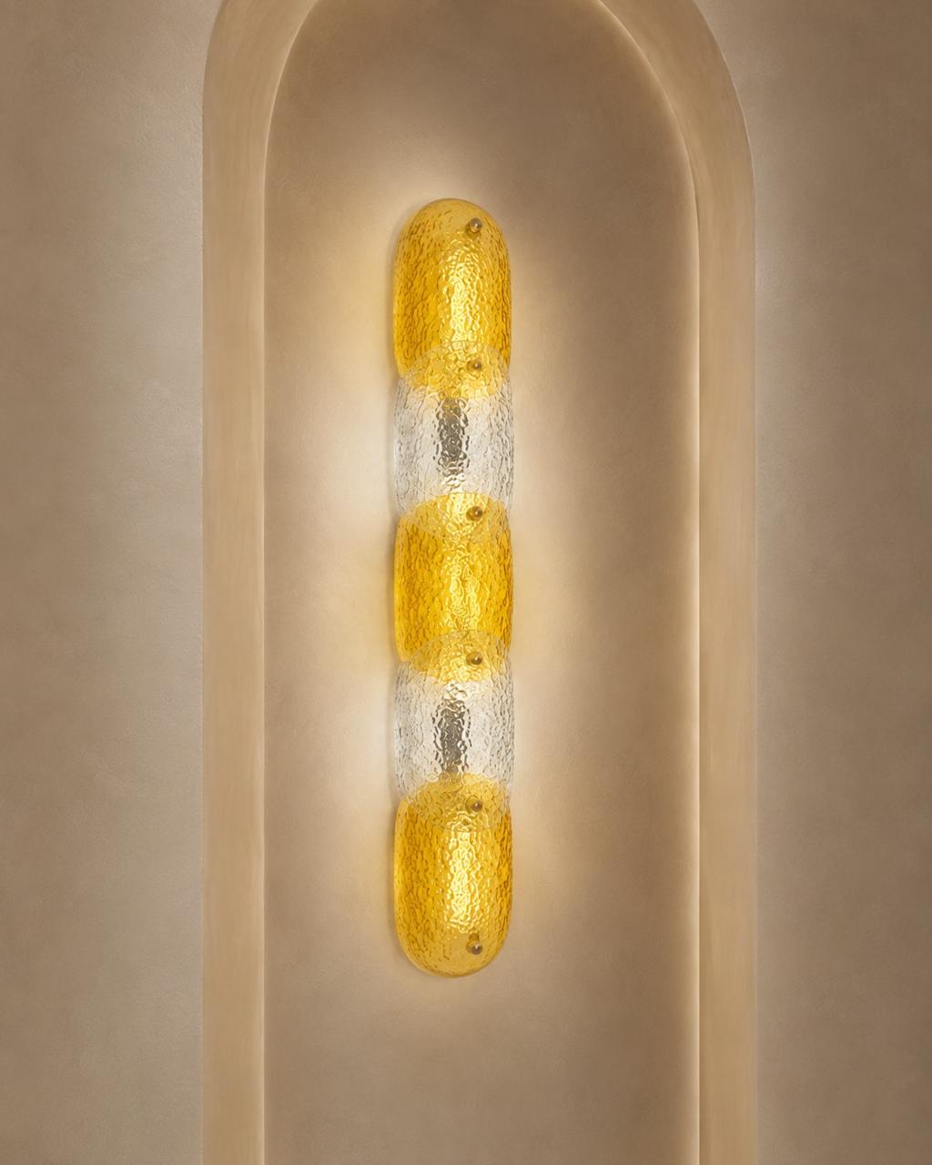 A modern vertical wall sconce with three textured yellow glass oval bulbs set in a clear glass column, mounted on a beige plaster wall with an arched recess.