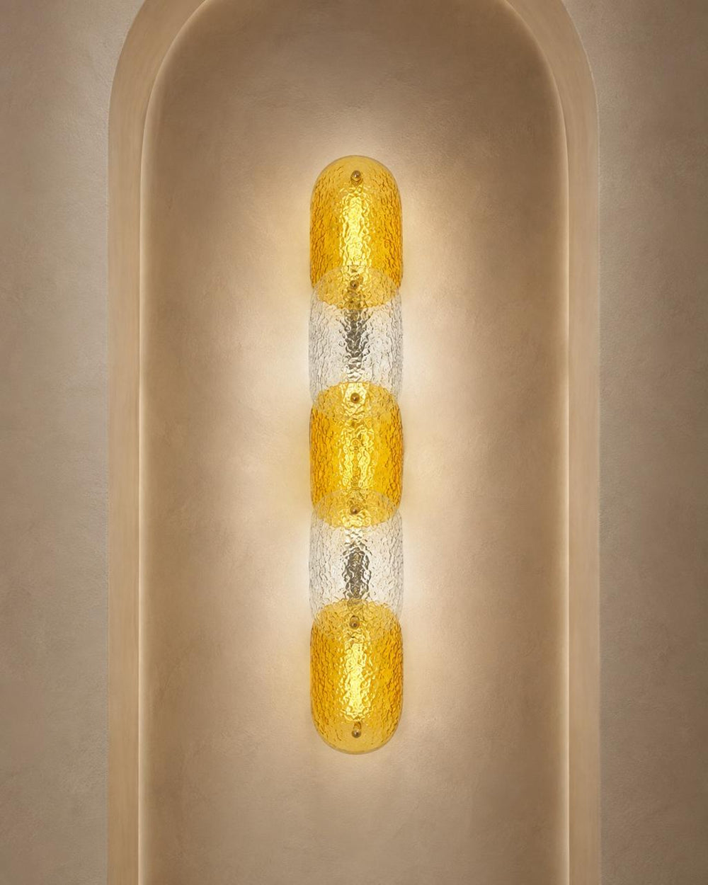 A modern vertical wall sconce with three textured amber-yellow glass oval shades separated by clear textured glass, mounted on a beige plaster wall in an arched niche.