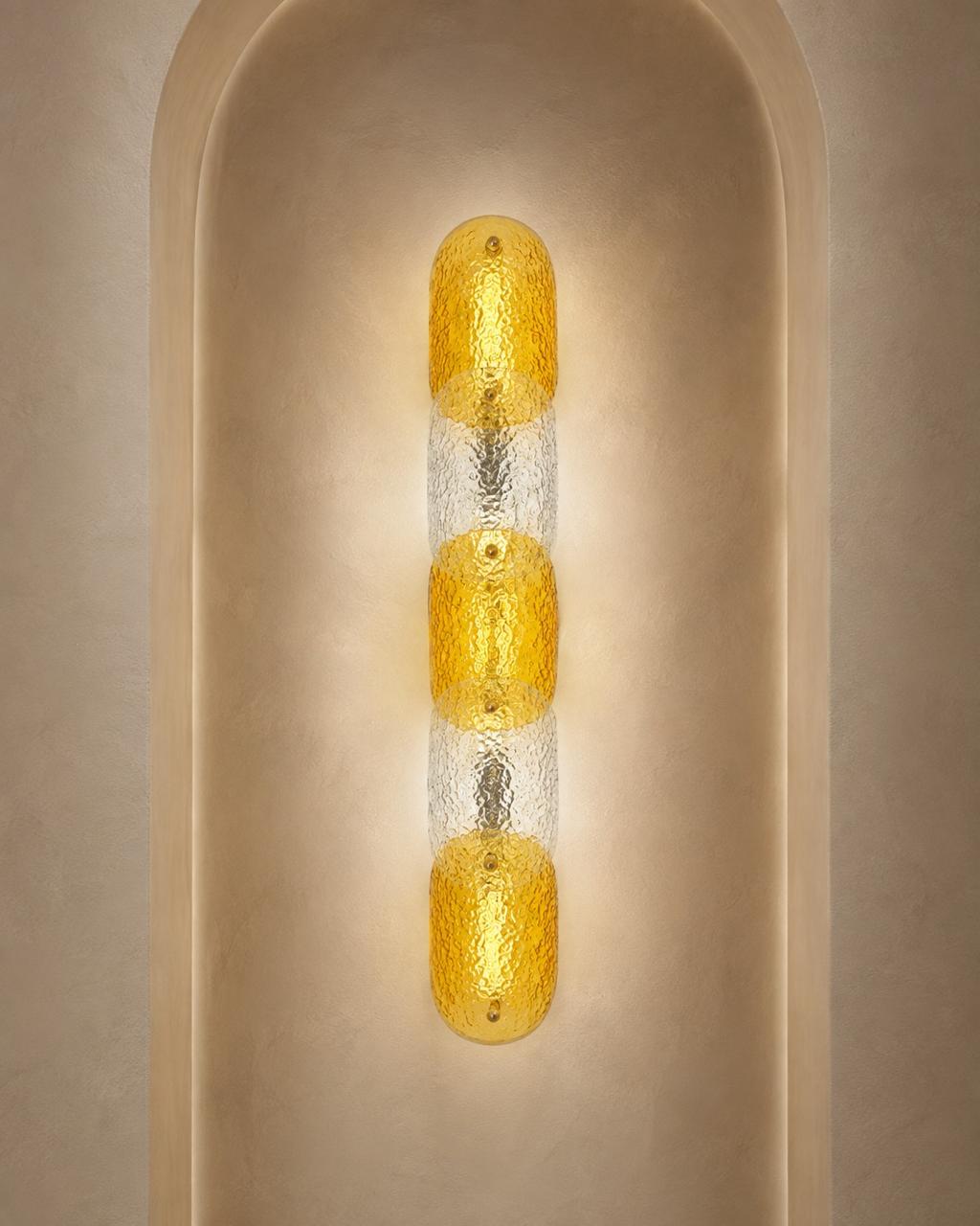 A modern vertical wall sconce with three textured amber-yellow glass oval shades separated by clear textured glass, mounted on a beige plaster wall in an arched niche.