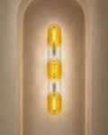 A modern vertical wall sconce with three textured amber-yellow glass oval shades separated by clear textured glass, mounted on a beige plaster wall in an arched niche.