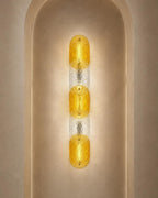 A modern vertical wall sconce with three textured amber-yellow glass oval shades separated by clear textured glass, mounted on a beige plaster wall in an arched niche.