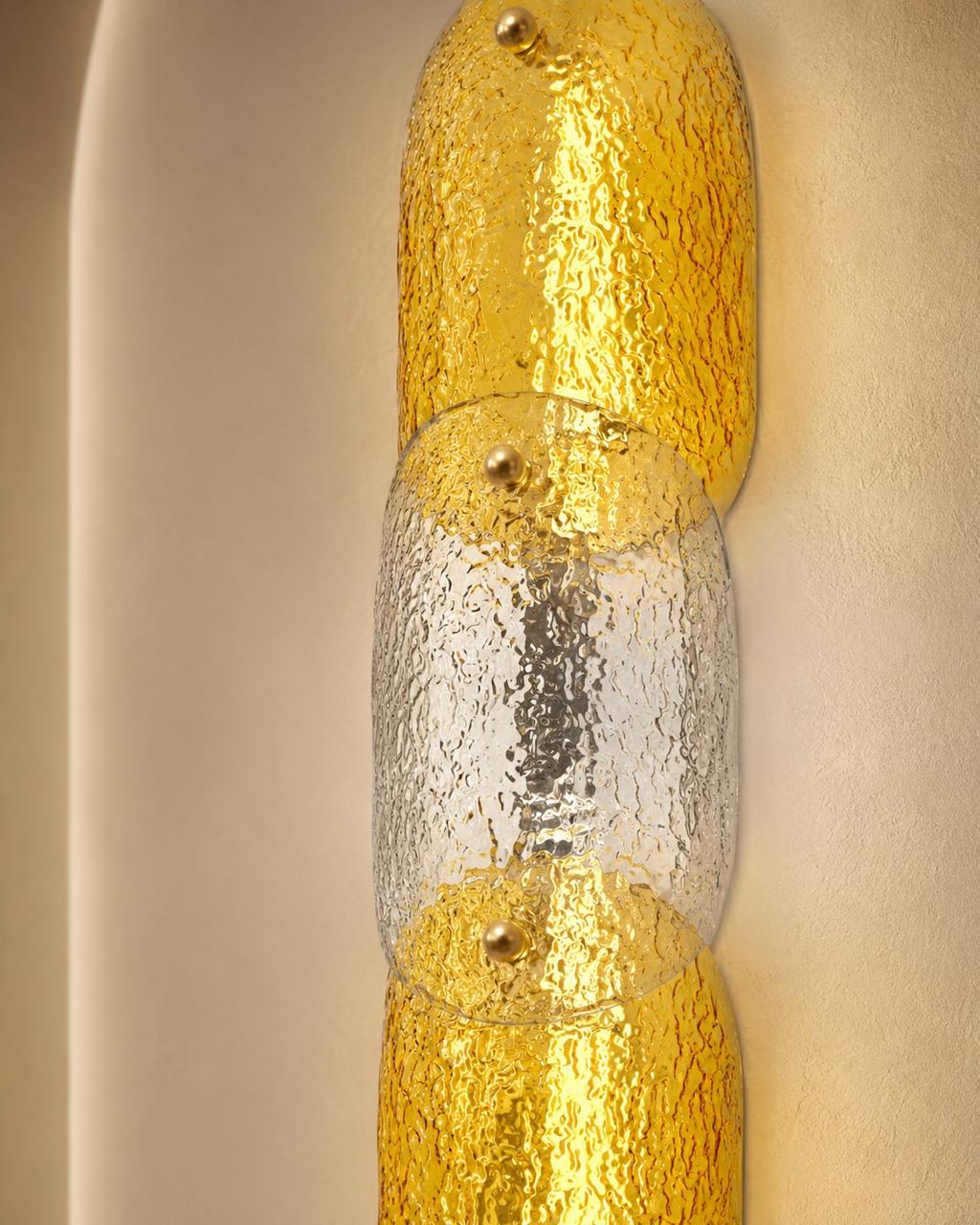 A close-up of a modern wall sconce featuring textured gold and clear textured glass cylindrical segments, mounted on a warm beige textured wall.