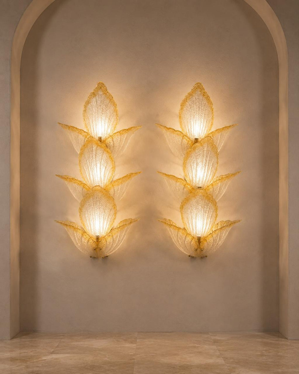 Two matching sculptural wall sconces mounted on a beige plaster wall within an arched alcove. Each sconce has a stacked, lotus-like form made of translucent textured glass or resin, glowing with warm light.