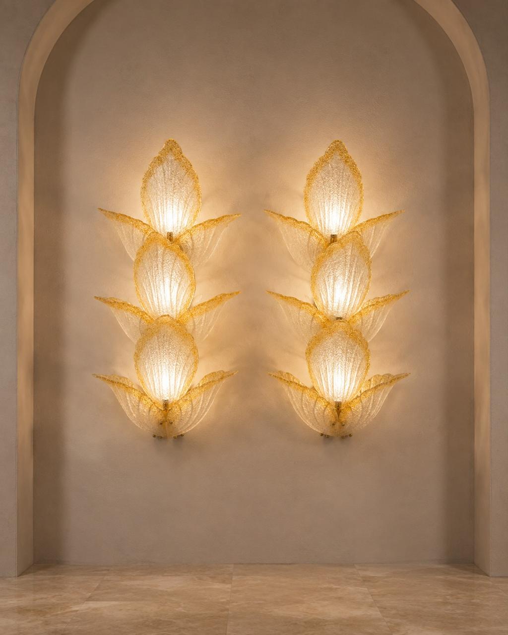 Two matching sculptural wall sconces mounted on a beige plaster wall within an arched alcove. Each sconce has a stacked, lotus-like form made of translucent textured glass or resin, glowing with warm light.