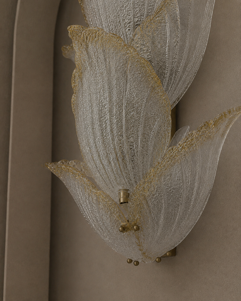A close-up of a wall-mounted decorative light fixture with layered translucent white glass petal-like shapes outlined in gold, attached to a beige wall.