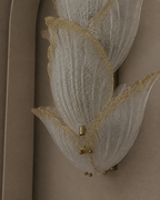 A close-up of a wall-mounted decorative light fixture with layered translucent white glass petal-like shapes outlined in gold, attached to a beige wall.