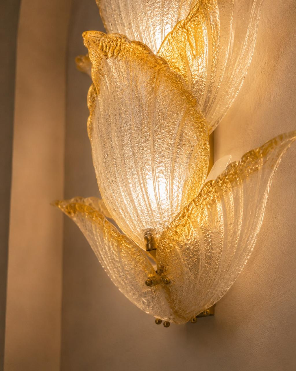 A close-up of a sculptural wall sconce with three overlapping translucent petal-like glass shades, glowing warm white from within and edged with metallic gold detailing, mounted on a beige wall.