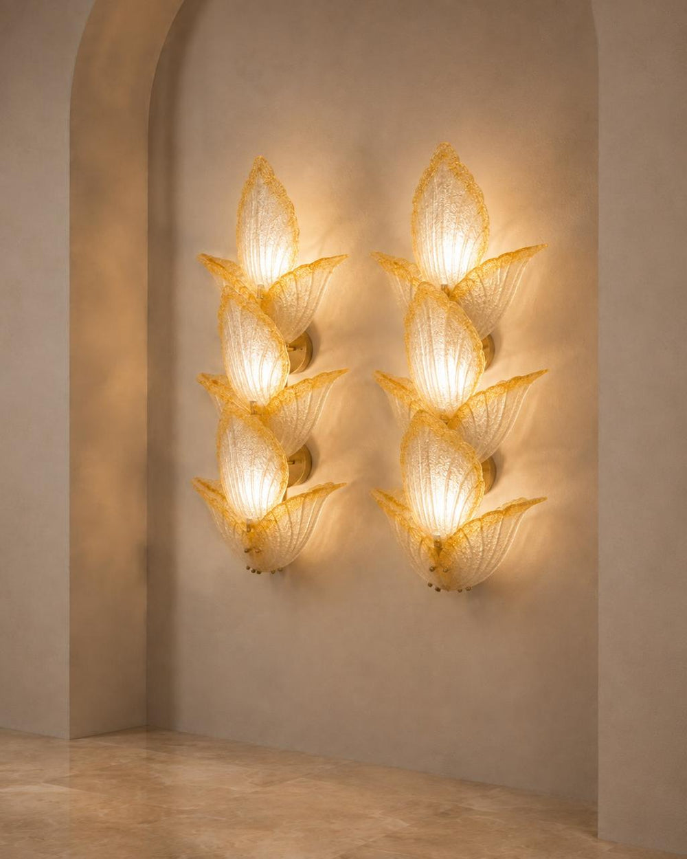 Two matching sculptural wall sconces mounted on a beige plaster wall in an arched alcove. Each sconce has a warm glowing organic, flower-like form made of translucent textured material with a gold-toned base.