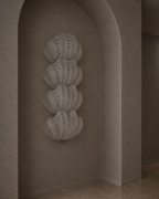 A textured, sculptural wall-mounted piece featuring four stacked, ribbed shell-like forms in a warm taupe-gray color, set within an arched alcove on a textured wall.