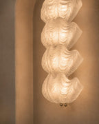 A modern wall-mounted light fixture with four stacked, organic, wavy shell-like forms in frosted white glass, glowing with warm light against a textured beige wall.