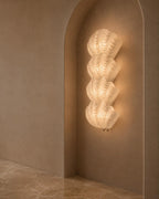 A sculptural, multi-tiered wavy wall sconce made of textured white glass, illuminated and mounted on a warm beige plaster wall within an arched niche.