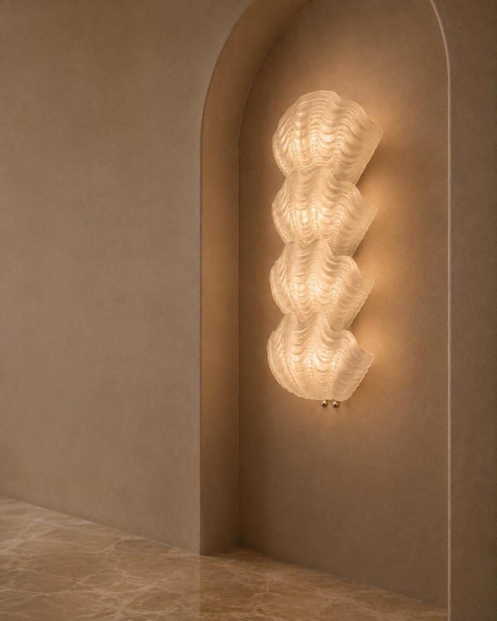A sculptural, multi-tiered wavy wall sconce made of textured white glass, illuminated and mounted on a warm beige plaster wall within an arched niche.