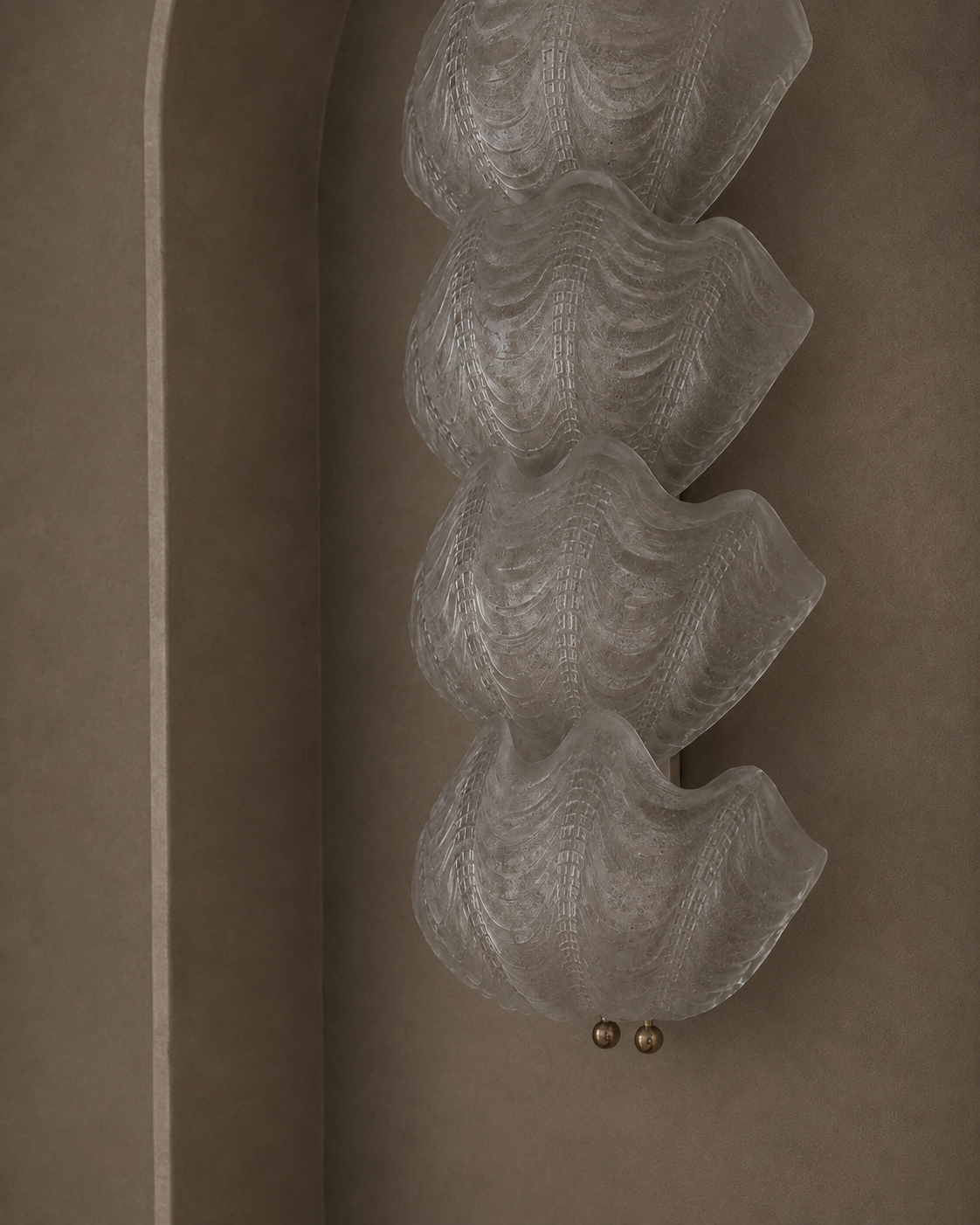 A sculptural wall sconce with four stacked, wavy translucent glass petals and two small gold finials, mounted on a warm taupe plaster wall in a recessed niche.