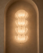 A sculptural, vertically stacked wall sconce made of layered, translucent ribbed glass, glowing with a warm light against a textured beige plaster wall in an arched niche.